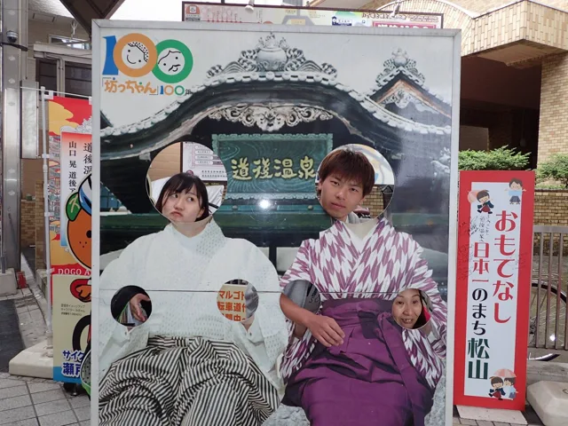 愛媛県松山市・道後温泉周辺（同ツアー写真から推定）での道後温泉周辺と見られる場所の顔出しパネルで撮影する参加者